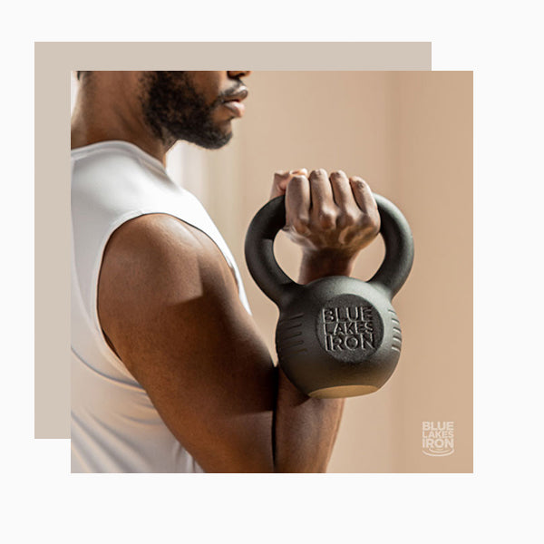 A man wearing a white sleeveless workout shirt does an arm curl with a 15lb kettlebell from Blue Lakes Iron.
