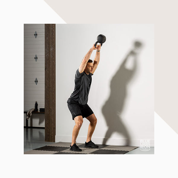 A man does an overhead two-handed kettlebell swing during a strength training workout in a home gym.