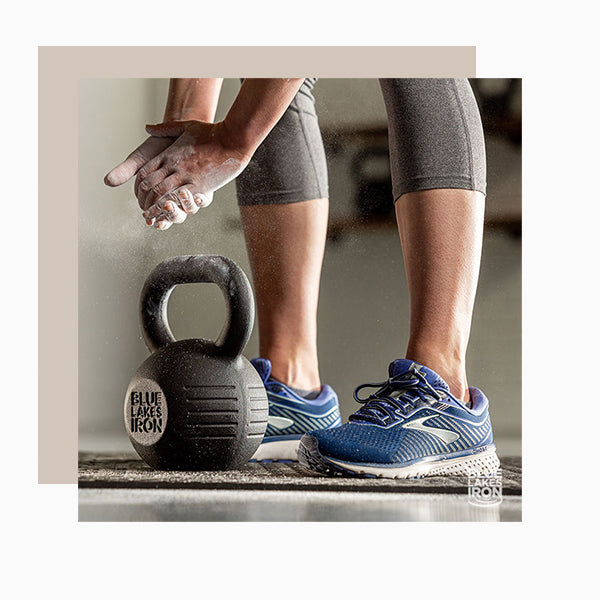 A person wearing blue tennis shoes bends over to pick up a cast iron kettlebell while putting lifting chalk on her hands.