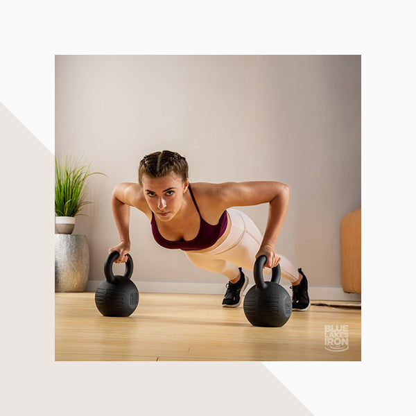 A woman is doing pushups using two Blue Lakes Iron kettlebells in a home gym to build strength.